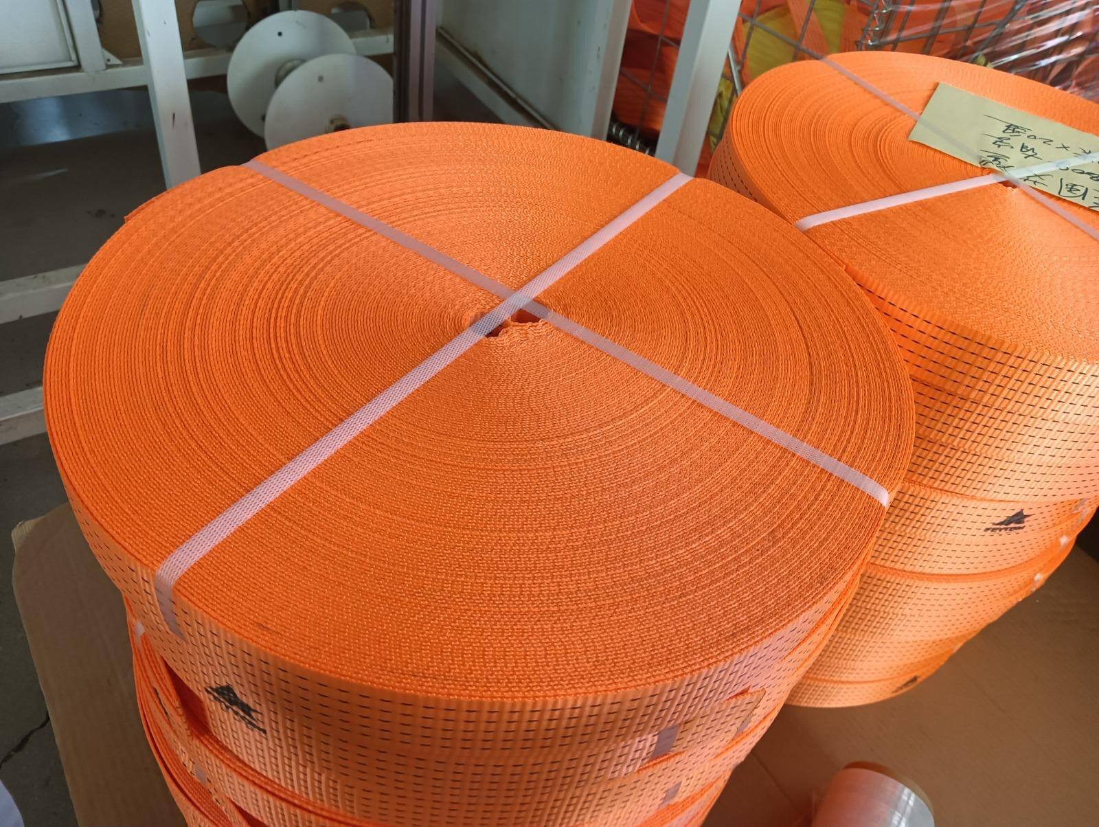 Large flat coils of orange lashing webbing, each banded crosswise with clear plastic strapping and stacked in a production area. A handwritten label sits on the coils behind.