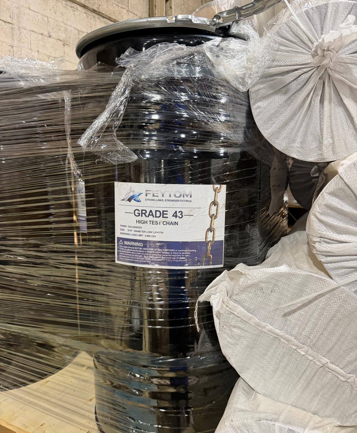 A black steel drum of Feytom Grade 43 high-test chain, shrink-wrapped on a warehouse pallet with white woven bulk bags stacked beside it. The drum carries a printed Feytom specification label.